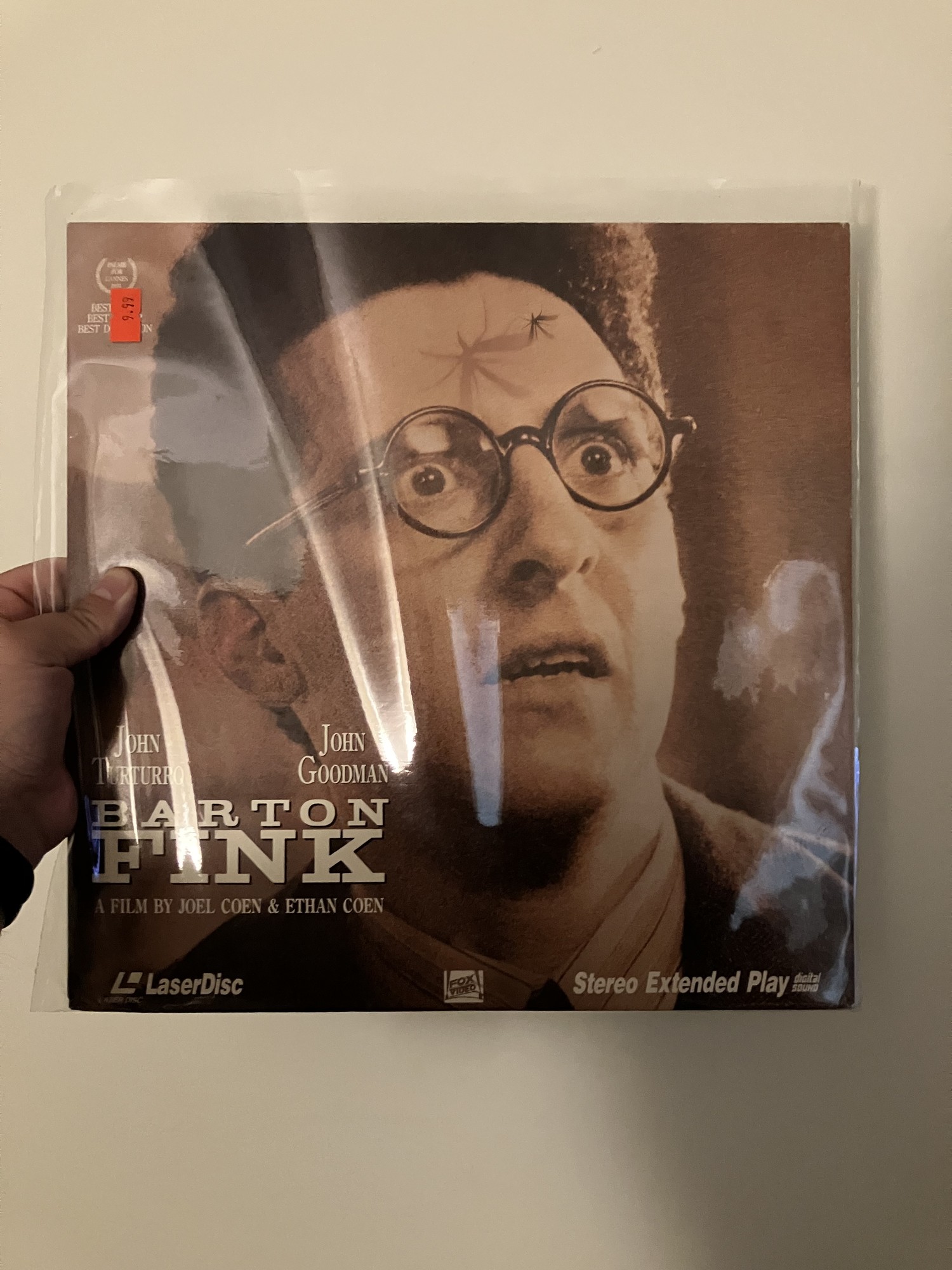 Alt-text: A person holds up a LaserDisc copy of the film Barton Fink in a clear plastic sleeve. The cover shows a close-up of John Turturro’s character, wide-eyed and wearing round glasses, with a bug crawling on his forehead. The title Barton Fink appears in bold white letters across the bottom, alongside the names John Turturro and John Goodman. The LaserDisc and 20th Century Fox logos are visible at the bottom, and there’s an orange price sticker in the top left corner marked $2.99.